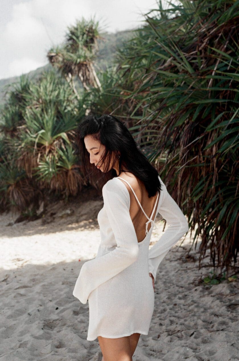 Back view of model wearing Cinnamon Swimwear off white Rayon Mini Dress, bikini coverup with long sleeves, elegant low cut back and back tie fasting's